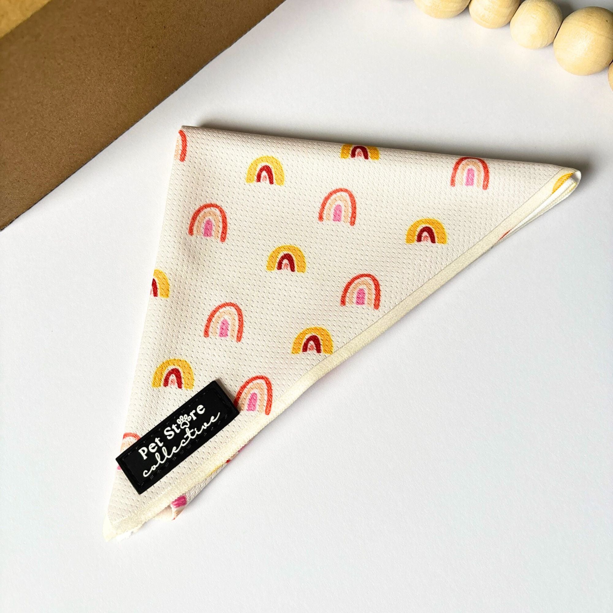 Front angled view of our Boho Rainbow Dog Bandana in soft golden yellow rainbow print, shown flat on a white background with natural wooden bead styling. Lightweight polyester tie-on design by Pet Store Collective.