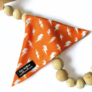 Angled view of our orange Thunderbolt Dog Bandana with white lightning bolts, tie-on design displayed on white background with wooden beads, stylish and comfortable for pets.