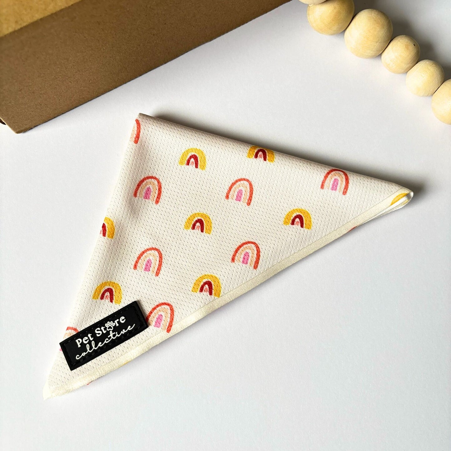 Angled top view of our Boho Rainbow Dog Bandana in warm yellow and peach rainbow design, displayed on a clean white background beside a cardboard box. Adjustable tie-on fit, lightweight fabric, designed in Australia by Pet Store Collective.