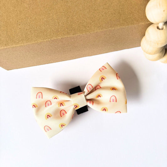 Our small Boho Rainbow Dog Bow Tie in golden tones with peach and yellow rainbow print, shown on white background with wooden bead decor. Lightweight and durable design with velcro strap that attaches easily to any pet collar.