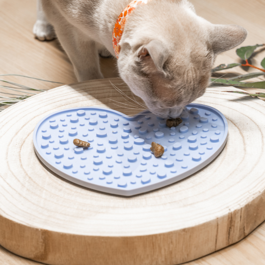 Our Sky Breeze Heart Licking Mat, measuring 20 cm by 17.4 cm, with a Burmese cat enjoying dry food from it. Suitable for both dry and wet pet food, this mat is perfect for cats and dogs. Made from 100% food-grade silicone, BPA-free, it features a subtle embossed Pet Store Collective logo on the back, promoting better digestion and mental stimulation with secure suction cups to keep it in place.