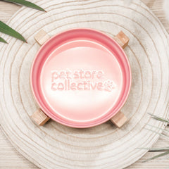 Petal pink ceramic pet bowl top view showing engraved Pet Store Collective logo with bamboo base on wooden board.
