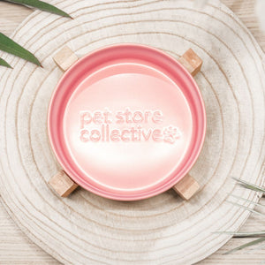 Petal pink ceramic pet bowl top view showing engraved Pet Store Collective logo with bamboo base on wooden board.