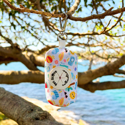 Our Summer Bliss Dog Poop Bag Holder hanging front-facing, showcasing its adorable beach print with palm trees, ice creams, sunnies, and sunshine tones. Made from durable quick-dry neoprene with a secure zipper holder and custom rubber dispenser, our holder keeps walks stylish and practical. Easily clips to your dog’s lead or handbag perfect for summer adventures.