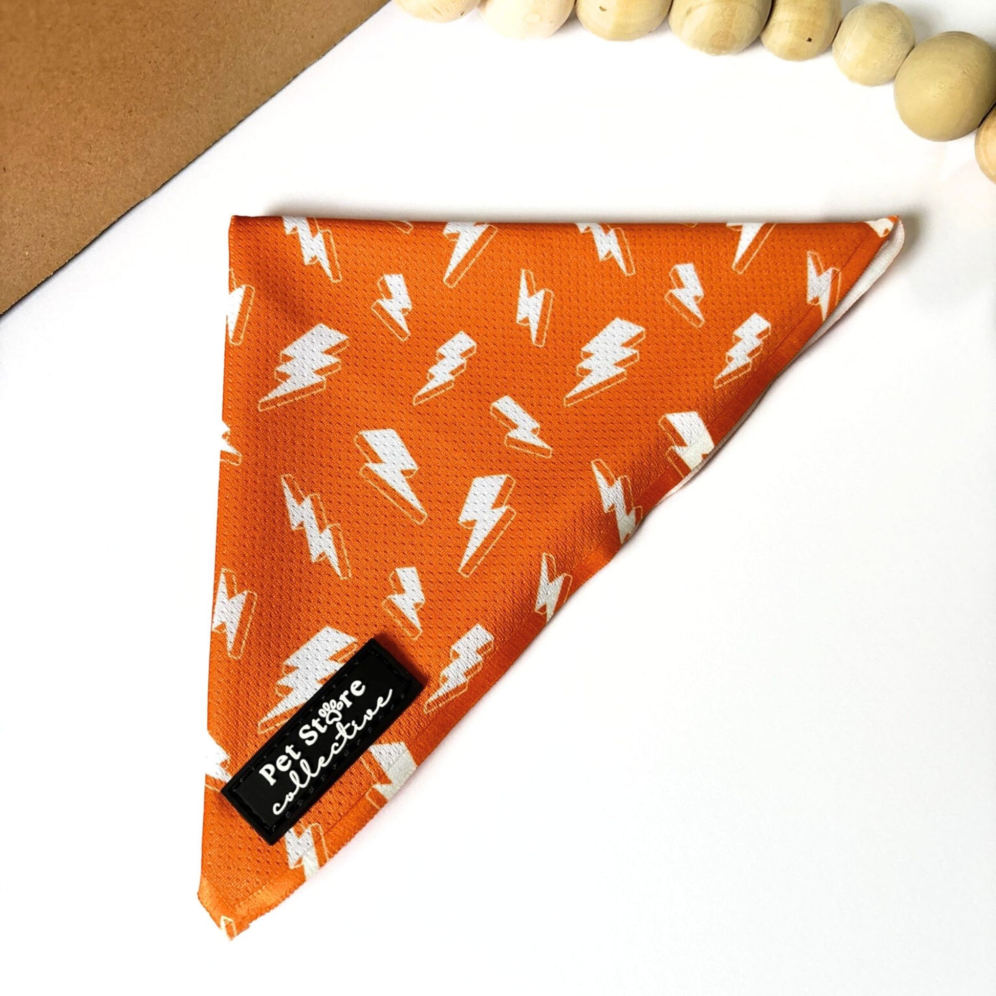 Front view of our Thunderbolt Dog Bandana in bright orange with white thunderbolts design. Shown flat with Pet Store Collective logo label, lightweight polyester fabric for dogs and cats.
