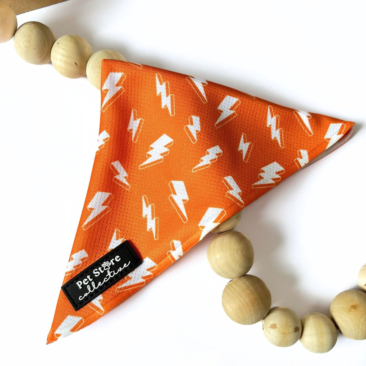 Angled view of our orange Thunderbolt Dog Bandana with white lightning bolts, tie-on design displayed on white background with wooden beads, stylish and comfortable for pets.