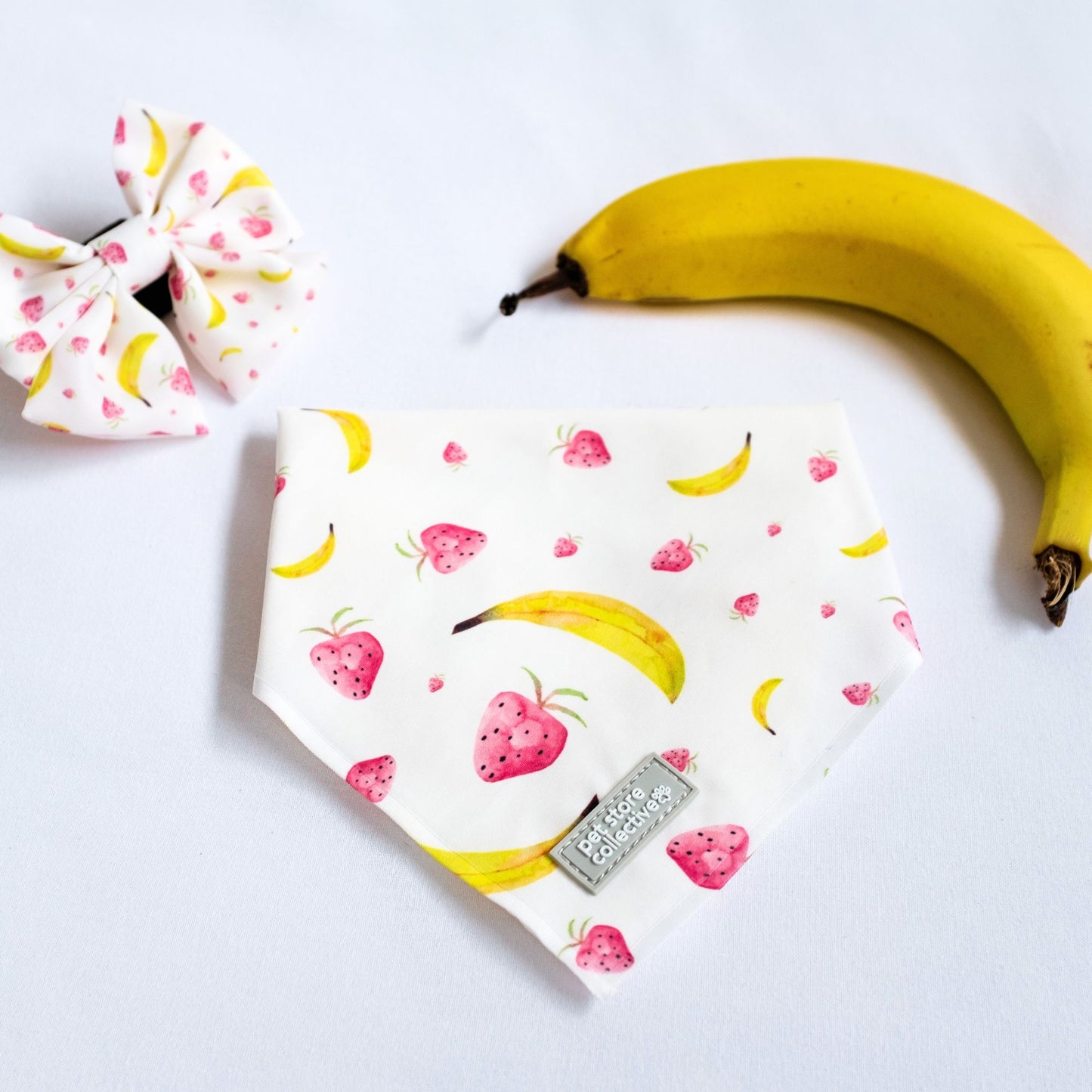 Close-up of our Tropical Pet Bandana and matching Sailor Bowtie in a vibrant strawberry and banana print, styled alongside a fresh banana for a fun summer vibe.